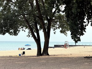 A beach with a large tree providing shade, a sandy area with beach chairs, and a calm sea in the background. - Chicago 1BR Near Lake Michigan & Pratt Beach – Free Parking (Chicago)