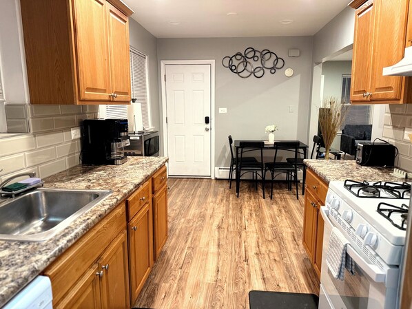 A kitchen with wooden cabinets, granite countertops, a gas stove, a microwave, and a dining area with a table and chairs. - Chicago 1BR Near Lake Michigan & Pratt Beach – Free Parking (Chicago)