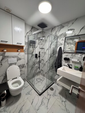 Un baño moderno con ducha de vidrio, inodoro blanco, lavamanos con espejo y paredes con diseño de mármol.