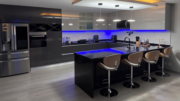 A modern kitchen with a central island, bar stools, stainless steel appliances, and blue under-cabinet lighting.