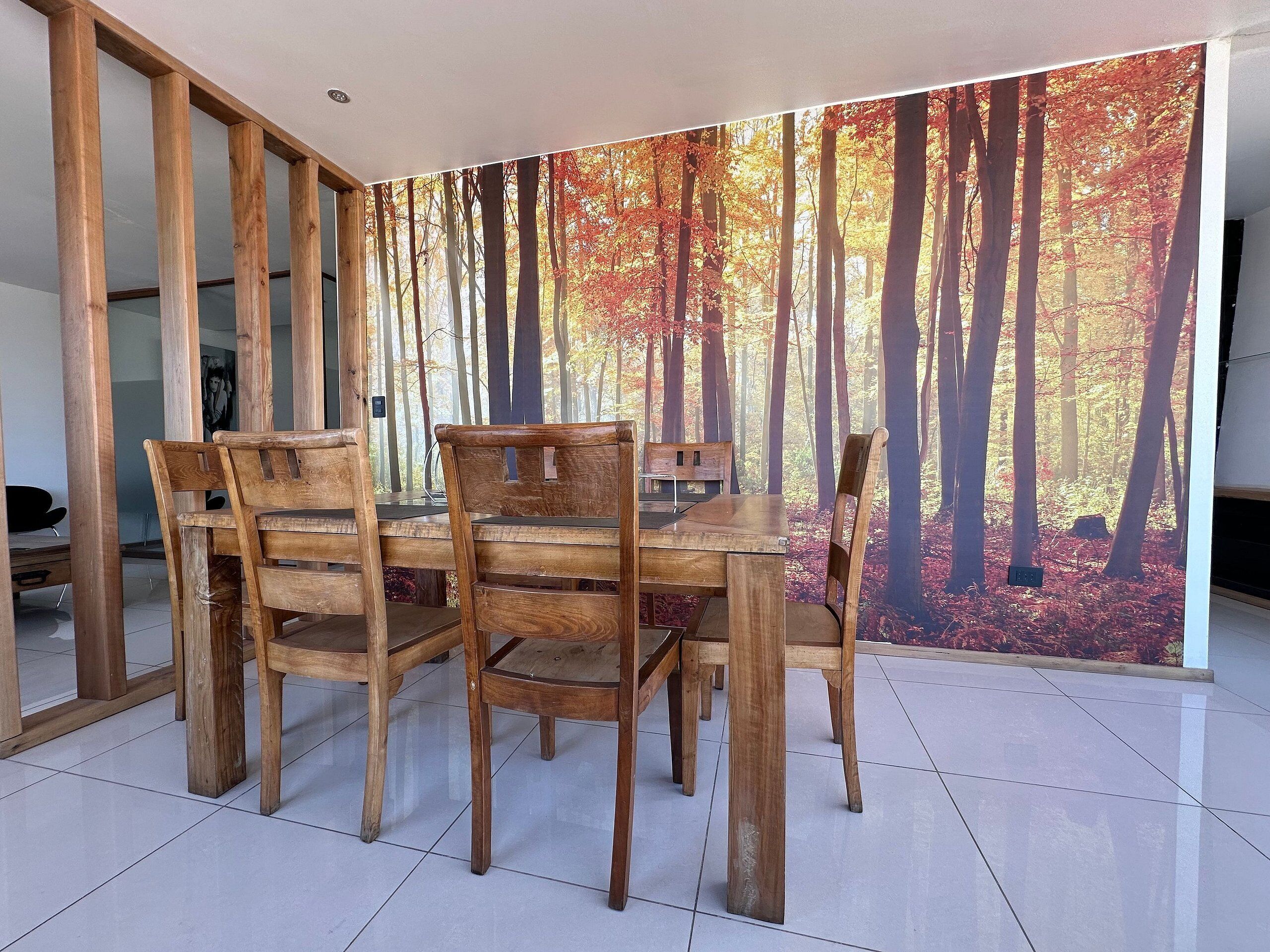 A dining area with a wooden table and chairs, a large forest mural on the wall, and a tiled floor.