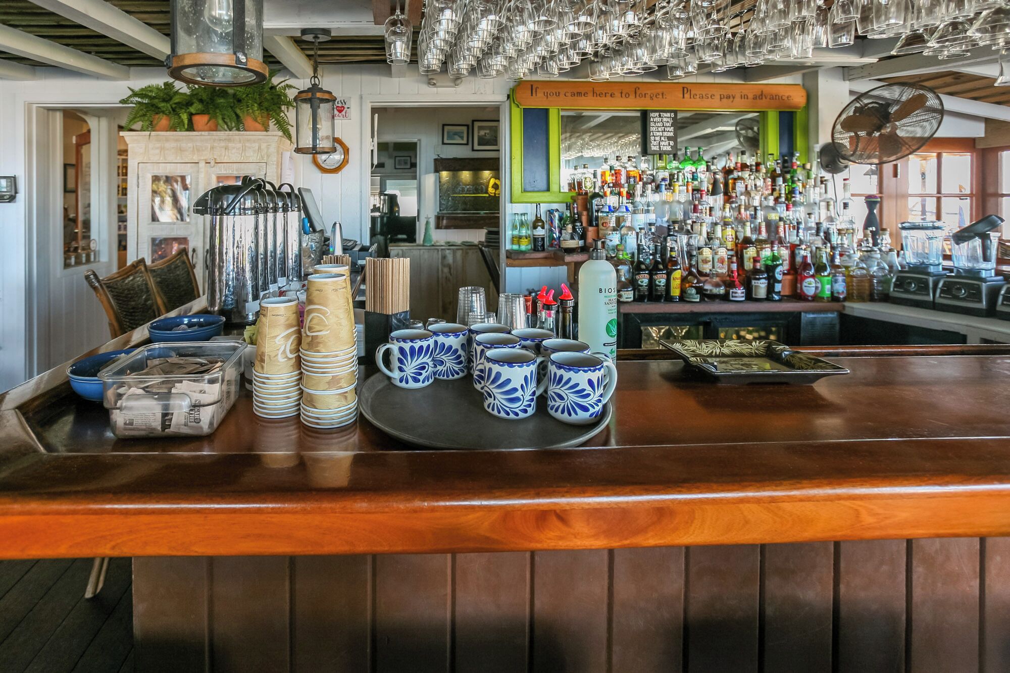 Un coin bar avec des piles de gobelets en papier, des tasses en céramique bleue et blanche, ainsi qu’une variété de bouteilles et de verres.