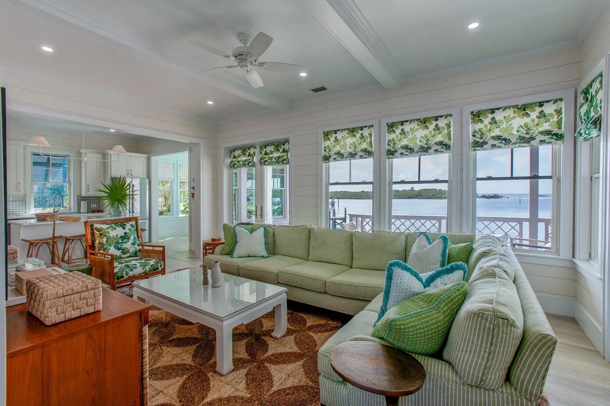 A spacious living room with a large window overlooking a body of water, a ceiling fan, and a dining area with wicker furniture.