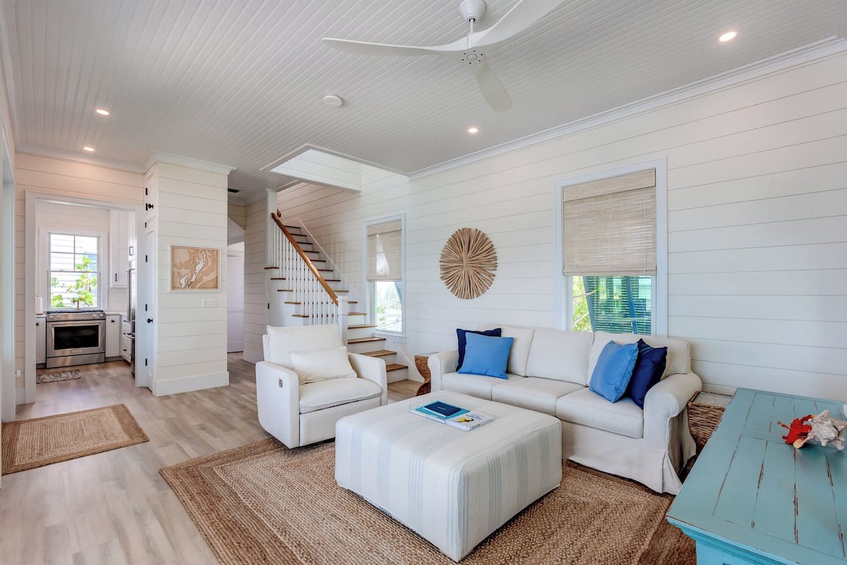A spacious living room with a white sofa, a coffee table, and a blue accent wall. A staircase leads to another level, and a kitchen is visible in the background.