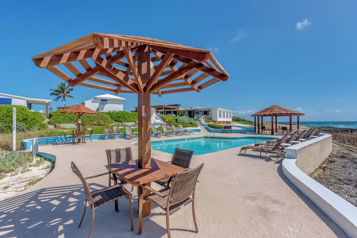 A poolside area with a wooden pergola, lounge chairs, and a table with chairs.