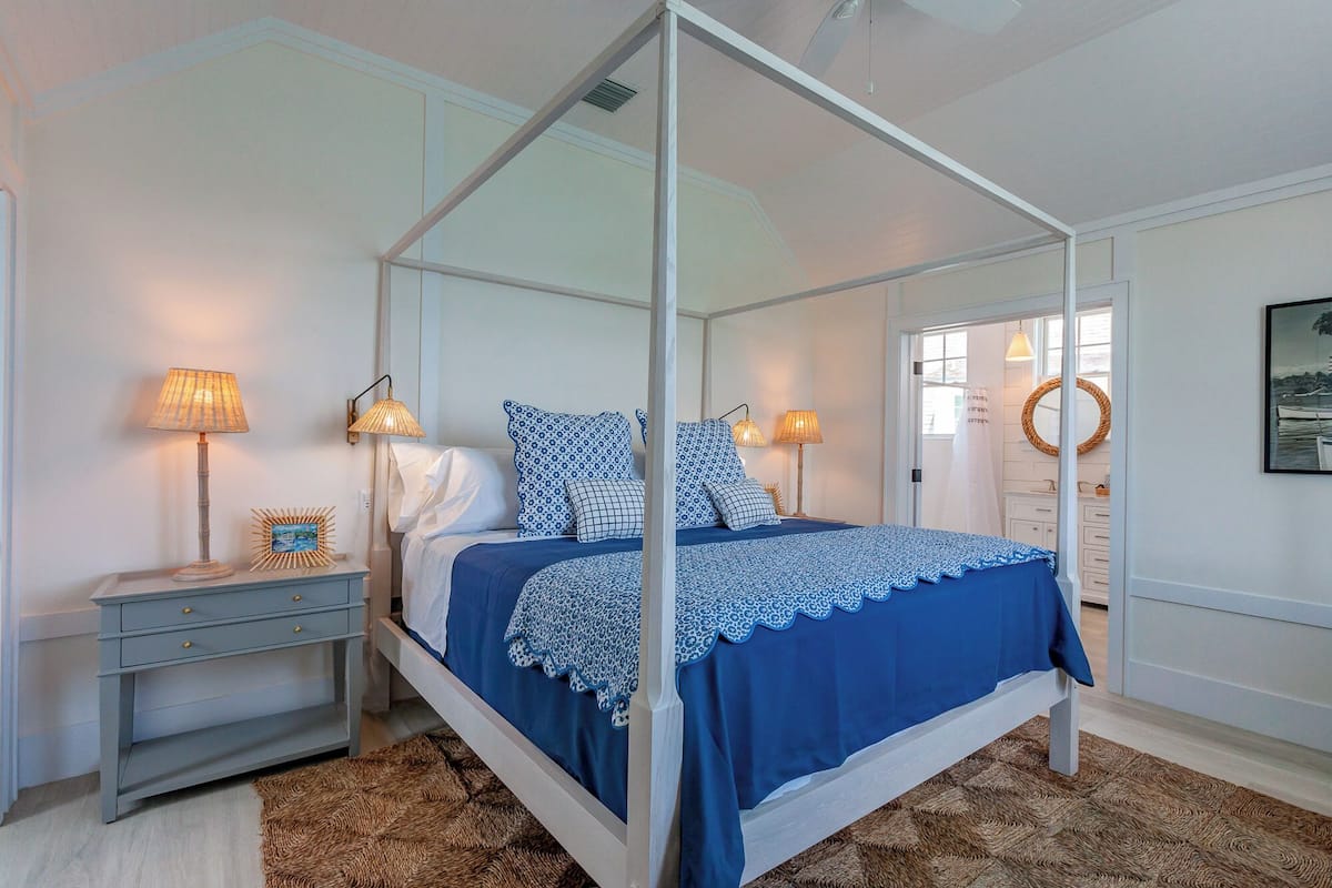 A four-poster bed with a blue bedspread, two bedside lamps, a nightstand with a decorative mirror, and a view of the outside through a window.