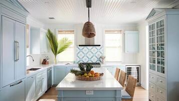 Una cocina con armarios azul claro, una isla central con fruta, un lámpara colgante y una ventana con vistas decorativas.