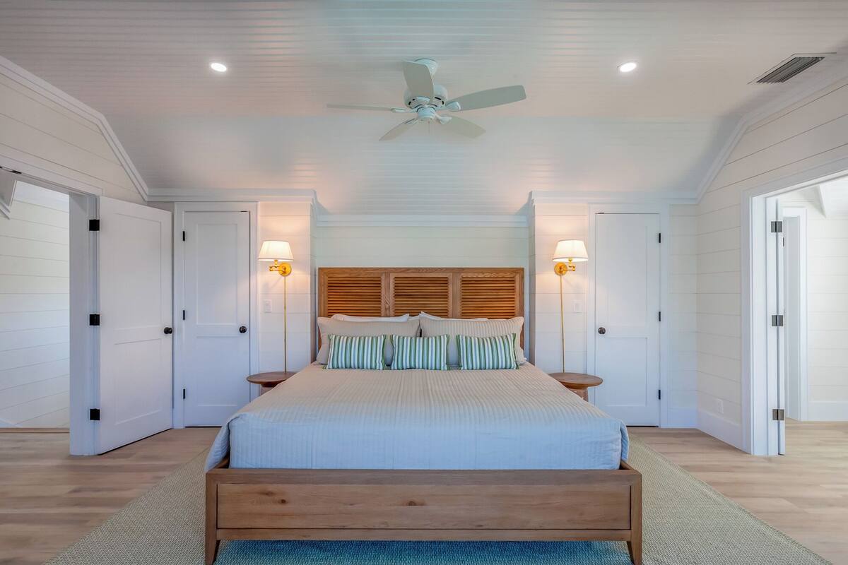 A bedroom with a large bed, wooden headboard, two bedside lamps, a ceiling fan, and white walls.