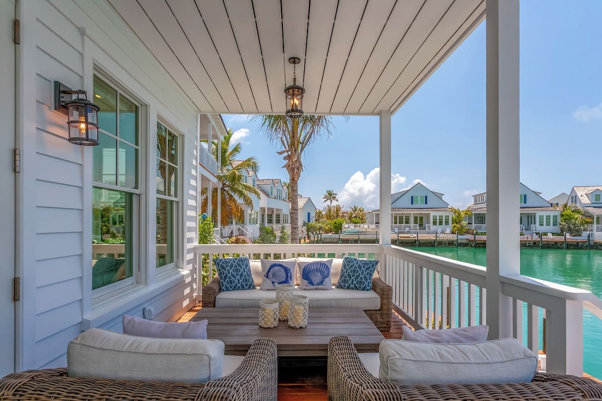 A covered porch with wicker furniture, a view of the water, and a row of houses in the background.