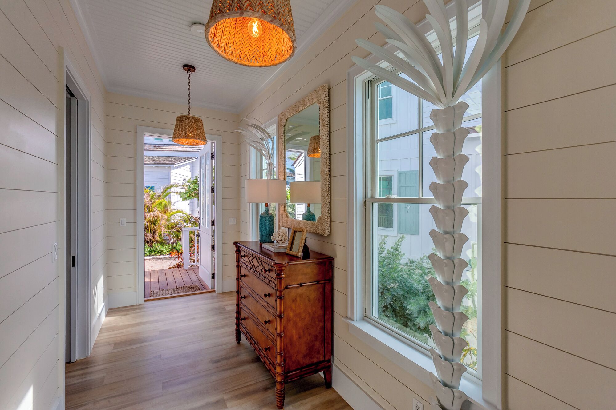 A hallway with wooden floors, a wooden dresser, a mirror, and a window with a decorative white curtain.