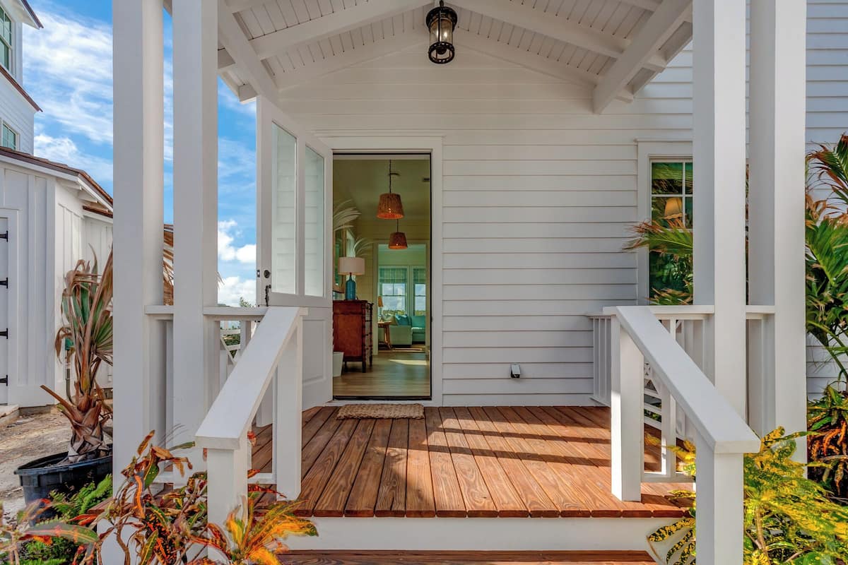 A covered porch with wooden decking and white railings, leading to a glass door into a room with a lamp and a view of the outdoors.