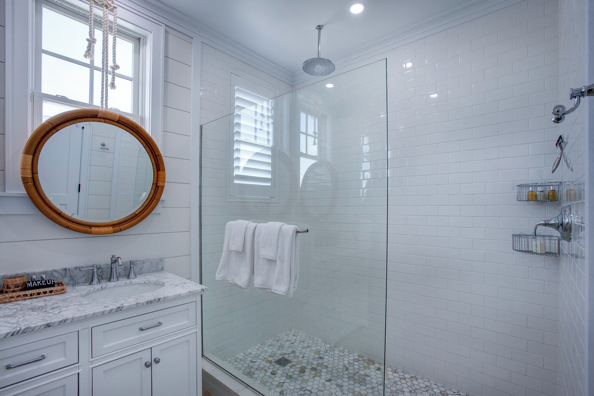 A modern bathroom with a glass-enclosed shower, white tiled walls, a marble sink, and a round mirror.