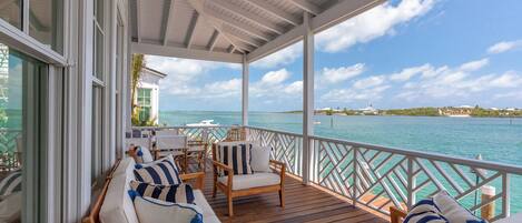 A covered outdoor seating area with wooden furniture and cushions, overlooking a turquoise sea and a distant island.