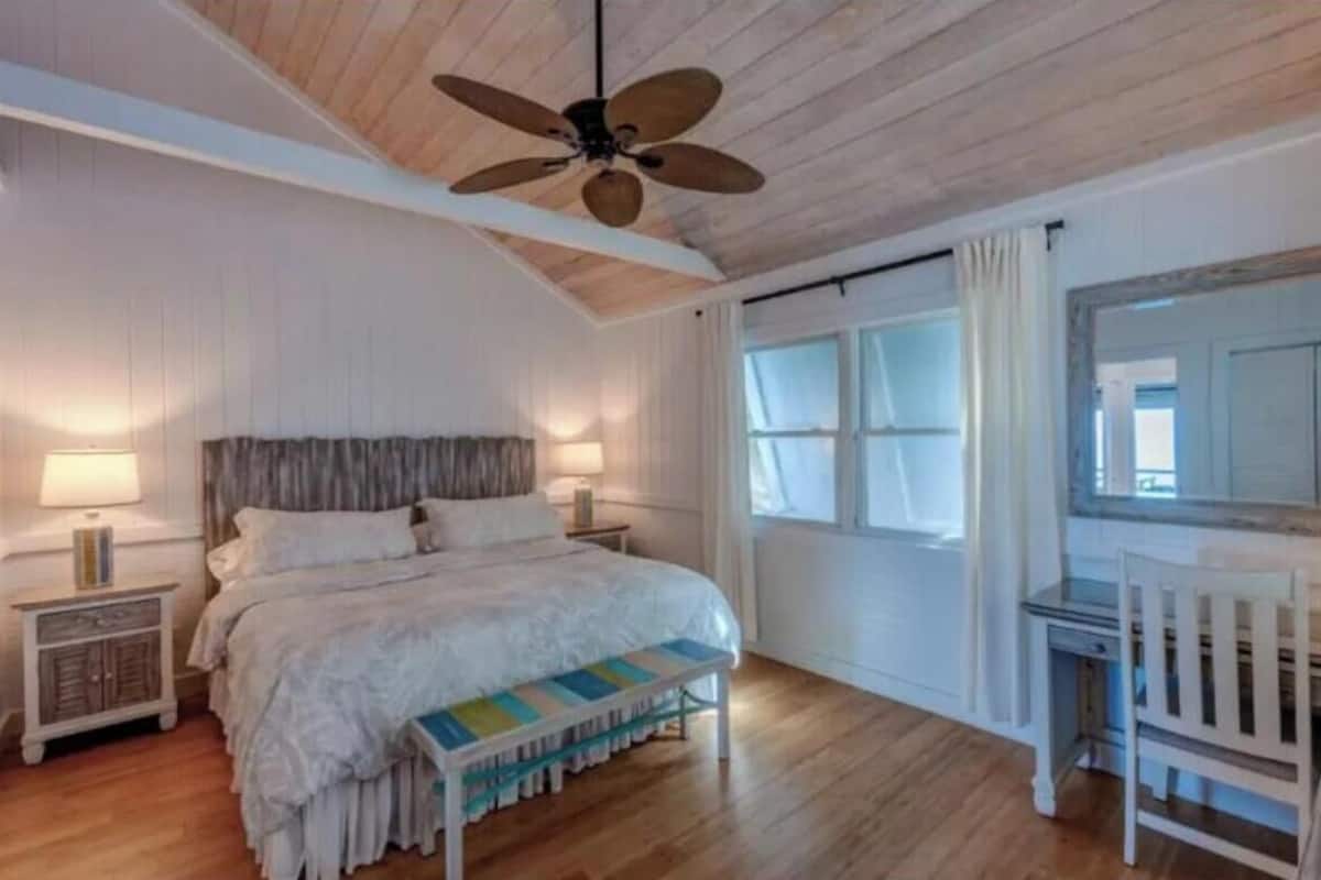 A bedroom with a wooden ceiling, a bed with white bedding, a nightstand with a lamp, a chair, and a window with curtains.