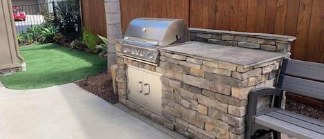 An outdoor barbecue grill with a stone base and a metal lid, positioned next to a wooden bench.