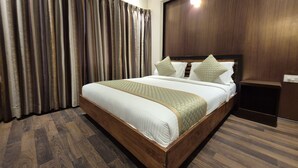 A neatly made bed with a wooden frame and a quilted bedspread, a wooden nightstand with a lamp, and a large window with curtains. - R D HOTELS HSR (Bengaluru)
