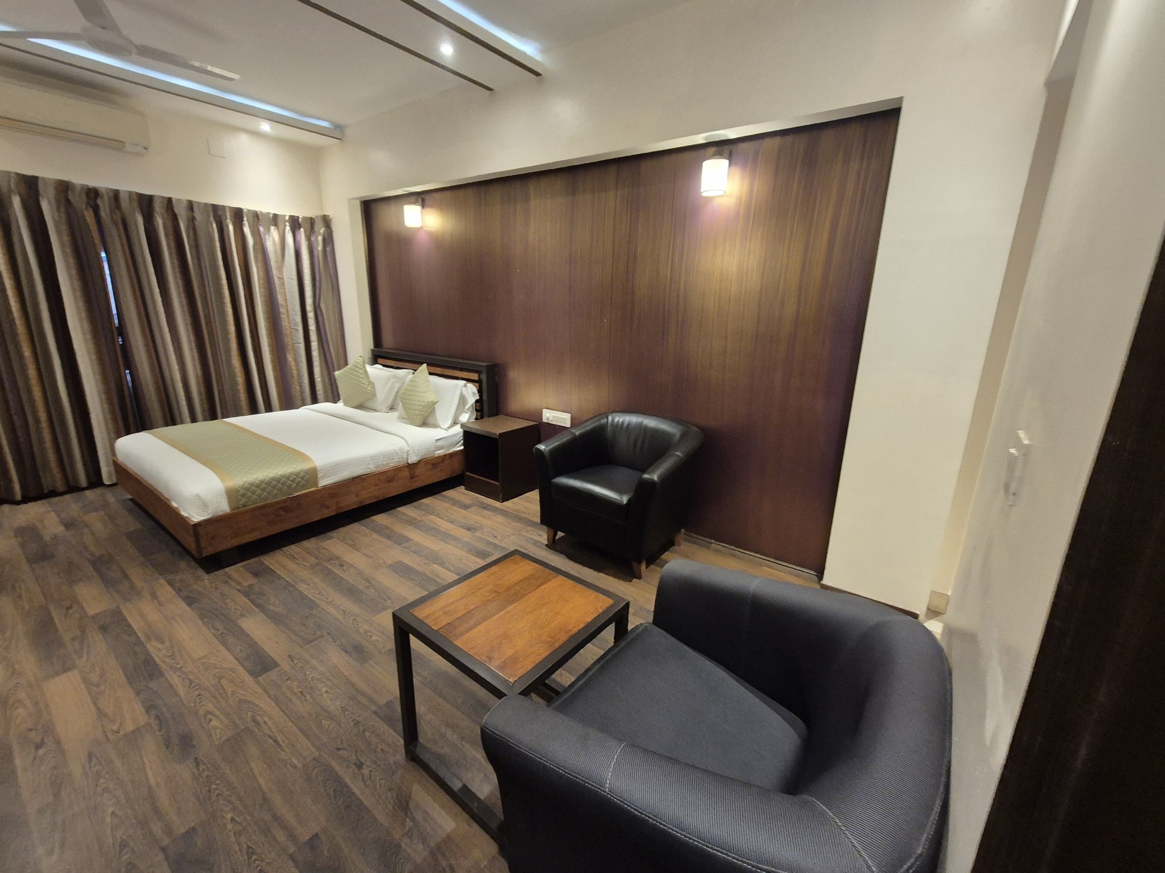 A modern hotel room with a wooden bed, a leather armchair, a small wooden table, and a dark wooden accent wall.