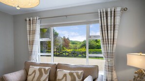 Ein Zimmer mit einem beigefarbenen Sofa, zwei gemusterten Kissen, einem Holztisch mit Lampe und Blick auf eine grüne Landschaft durch ein Fenster mit Karottenvorhängen.