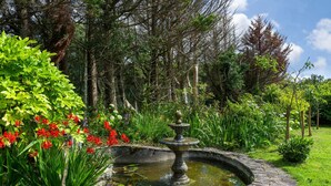 Ein Steinkunstbrunnen, umgeben von üppigem Grün und blühenden Pflanzen in einem Garten.