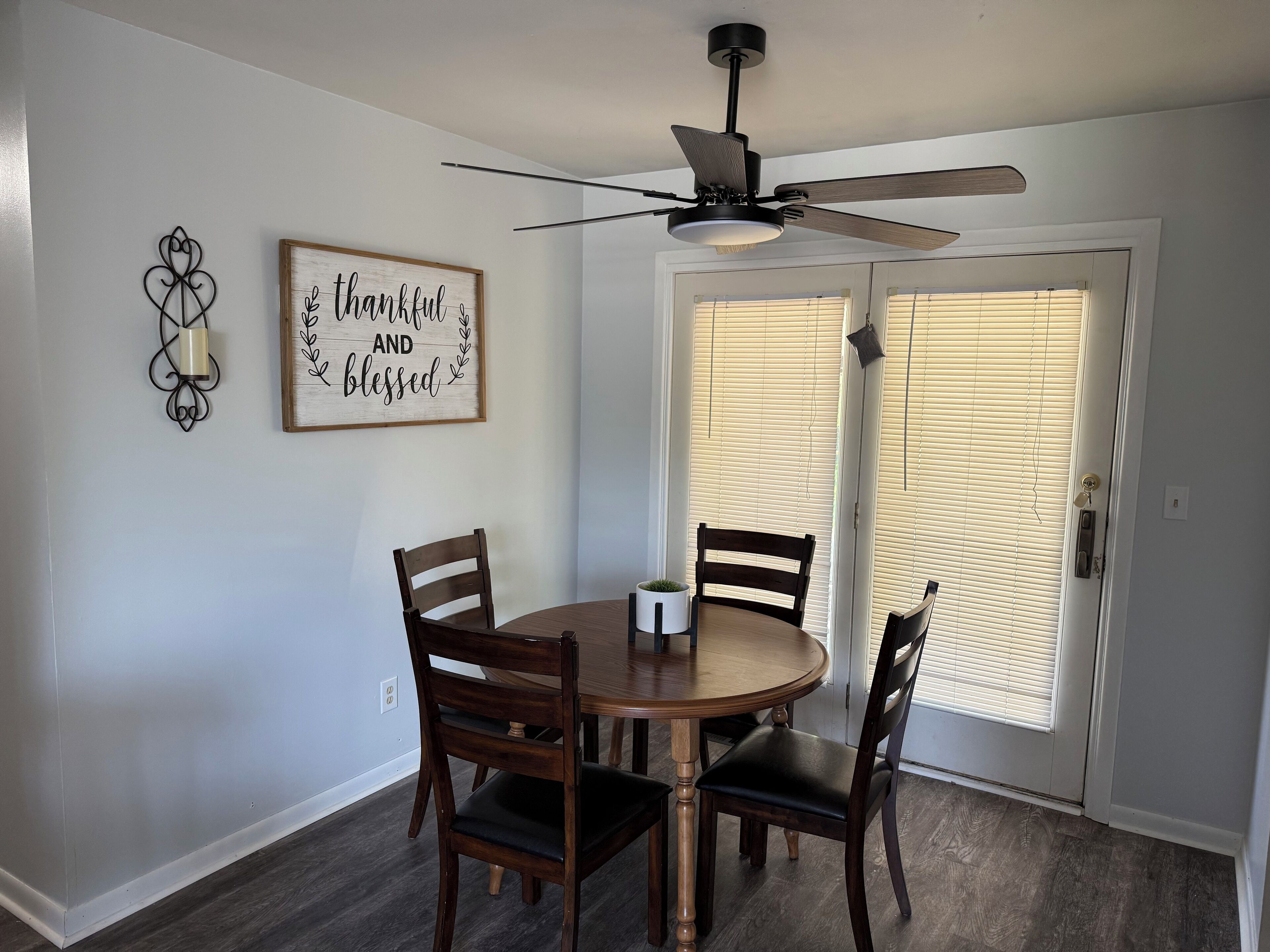 A dining area with a round table, four wooden chairs, a ceiling fan, and a door with blinds.