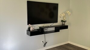 A room with a mounted TV on a black shelf, a power outlet, and a small potted orchid on the shelf. - Cozy 3-Bedroom Ranch style house with Huge Backyard & Fire pit (Indianapolis)