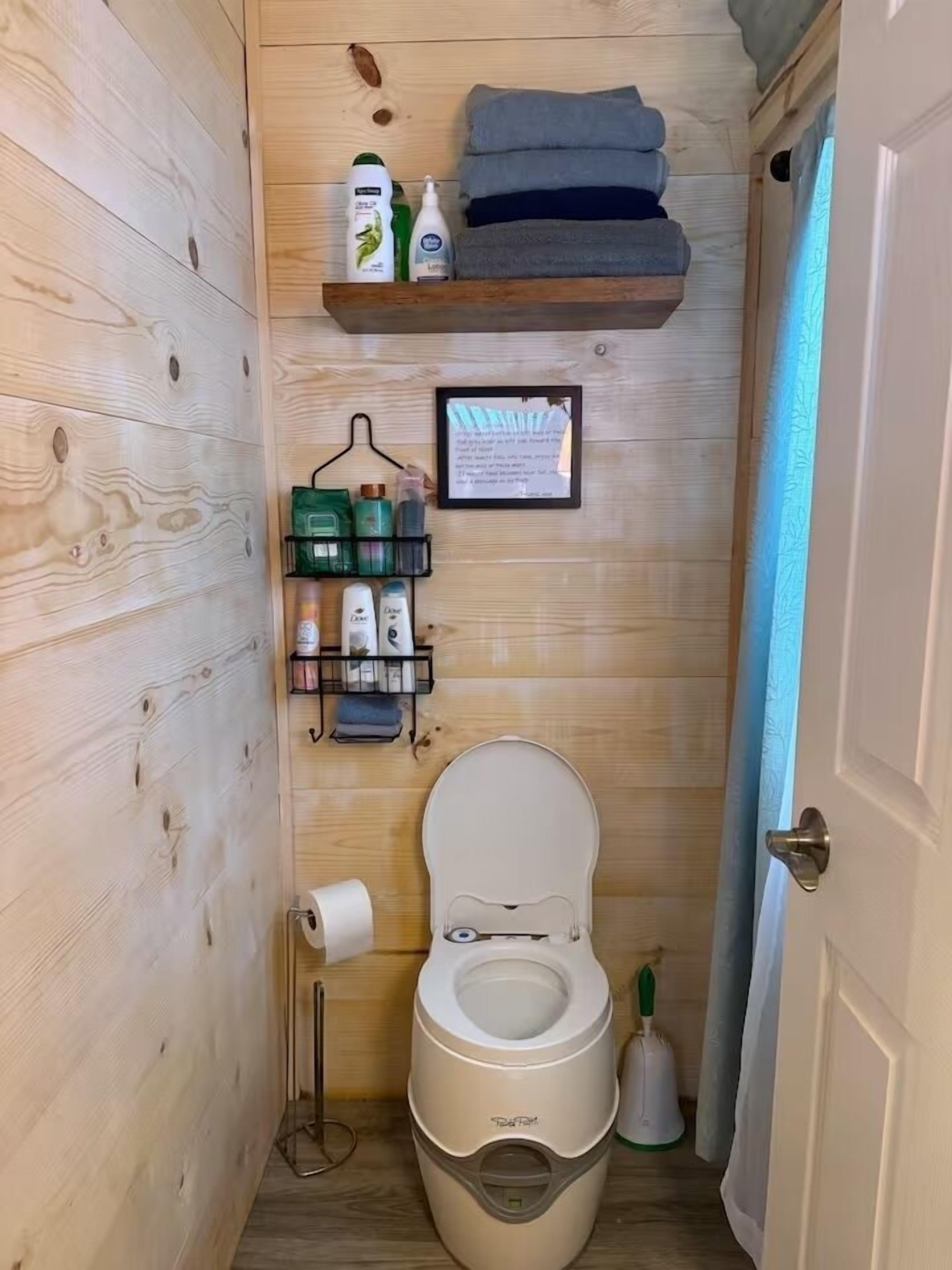 A compact bathroom with a toilet, a shelf with toiletries, a framed notice, and a broom.