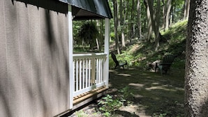 A small wooden cabin with a porch, surrounded by a forested area with benches and a dirt path.