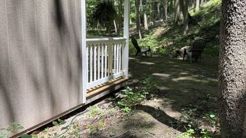 A small wooden cabin with a porch, surrounded by a forested area with benches and a dirt path.