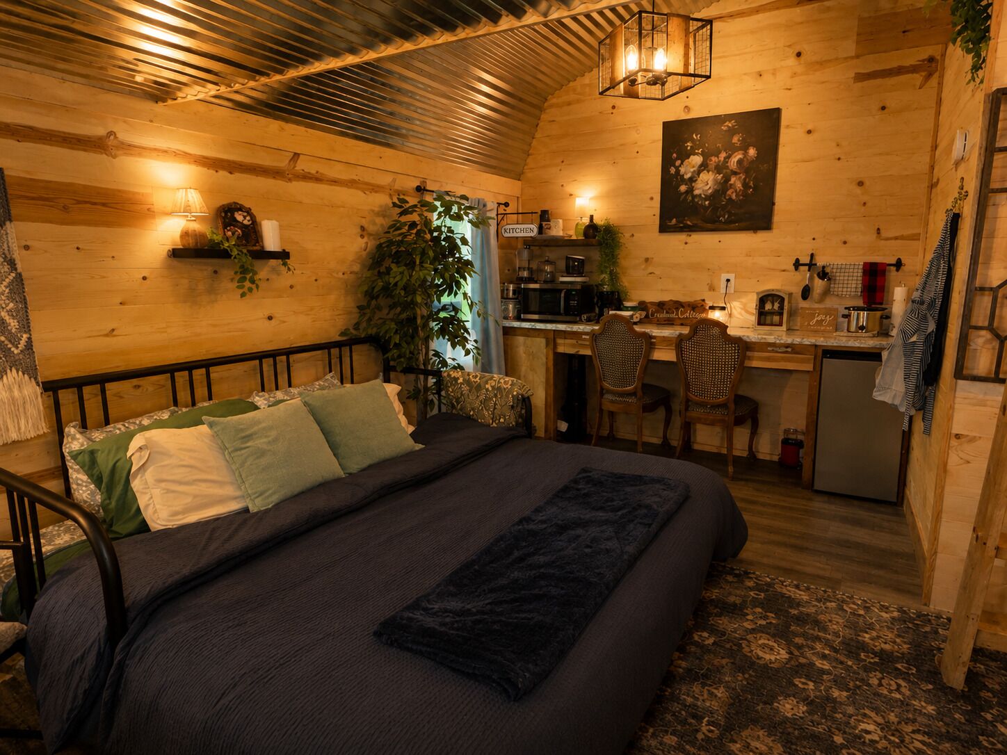 Acogedor dormitorio de cabaña de troncos con cama, mesita de noche, lámpara y una pequeña zona de escritorio con computadora, silla y electrodomésticos de cocina.