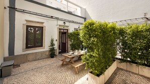 Une cour avec une table en bois et des bancs, entourée de verdure et bordée d’un bâtiment blanc doté d’une porte et d’une fenêtre.