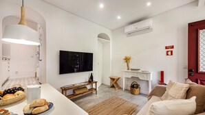 Un salon moderne avec un canapé blanc, une table basse, un téléviseur et un coin repas agrémenté d’un panier de fruits.
