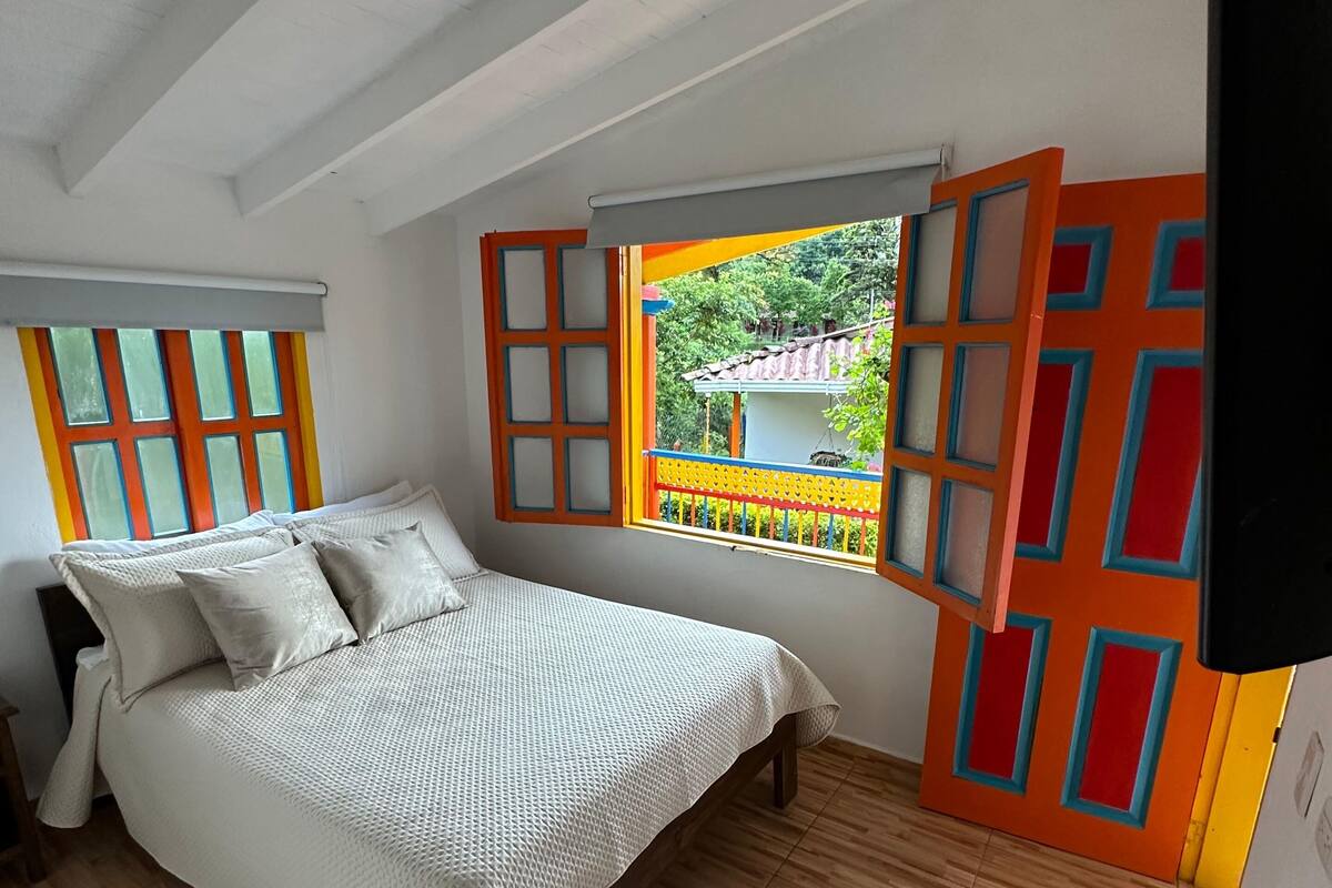 Una camera da letto con un letto, pavimento in legno, una finestra con porta aperta e vista su vegetazione.