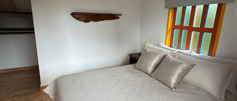 A bedroom with a large bed, wooden flooring, a window with colorful panes, and a wall-mounted shelf.