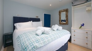 A neatly made bed with white linens and a blue patterned bedspread, a dark blue headboard, a nightstand with a lamp, a white dresser with a mirror, and a blue door in the background.