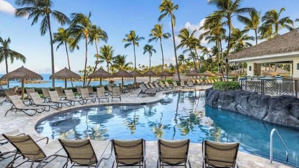 A resort pool area with lounge chairs, palm trees, and a thatched-roof structure. - View Stunning Sunsets in this Ocean View 1 Br @ MVC Ko Olina Beach Club (Kapolei)