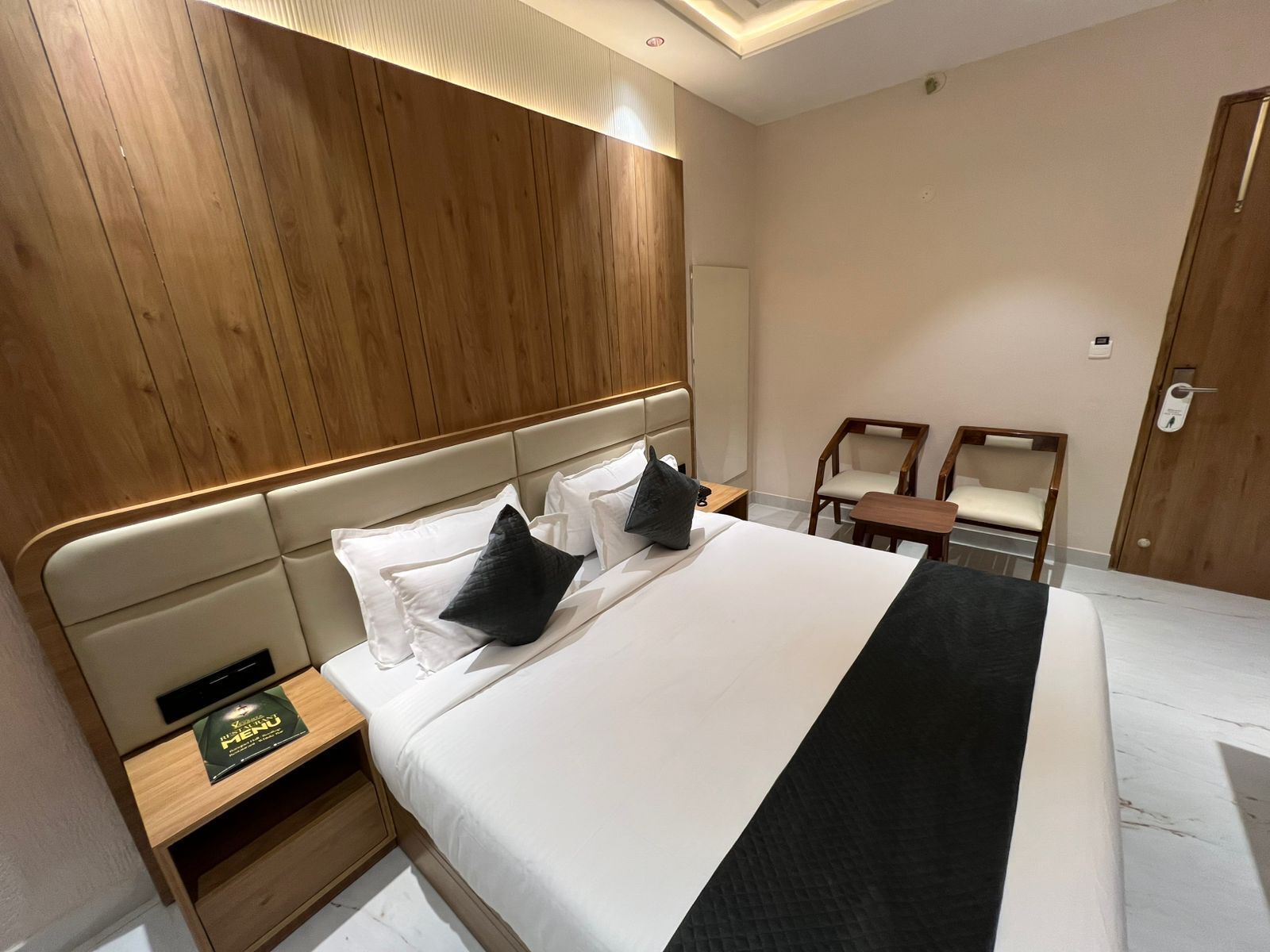 A hotel room with a large bed, wooden headboard, two bedside tables, a nightstand, a small wooden table, two chairs, and a door with a handle.