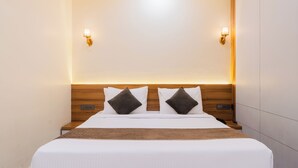 Una cama bien tendida con sábanas blancas y dos almohadas oscuras, cabecera de madera y dos lámparas fijadas a la pared.