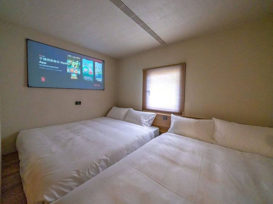Habitación de hotel con dos camas, un televisor con aplicaciones de streaming, una ventana con persianas y una pared lisa.