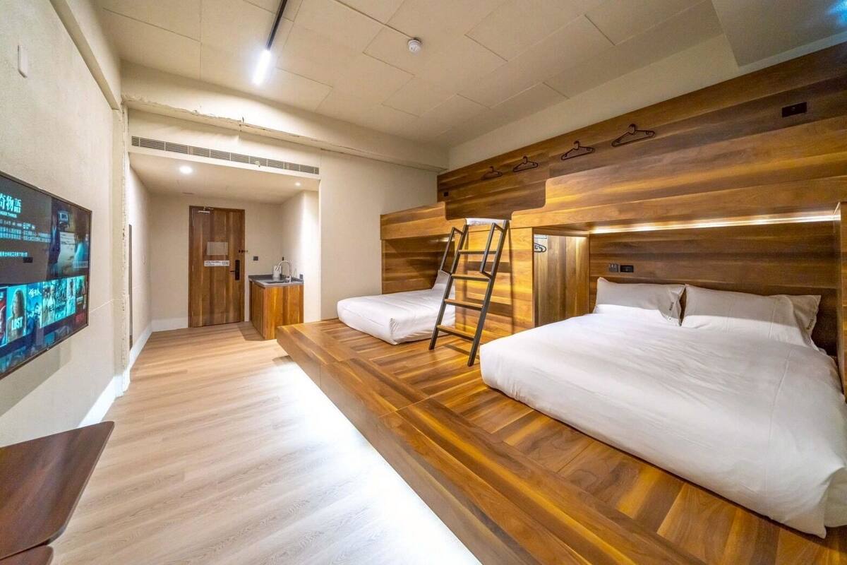 Una habitación de hotel con dos camas, piso de madera, televisor en la pared y baño con lavamanos y espejo.