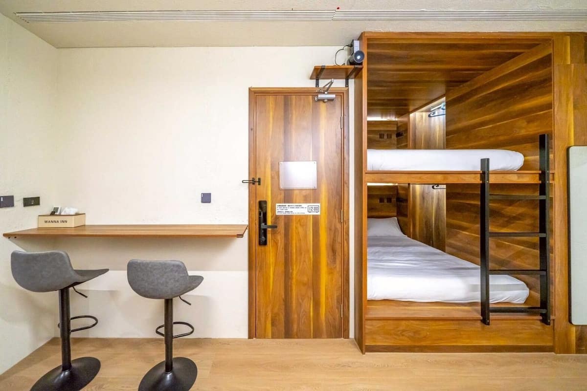 Una habitación con una cama litera, un escritorio, dos taburetes altos y una puerta de madera.