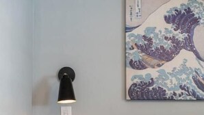Un dormitorio con una cama, una mesita de noche, una lámpara de pared y una pintura japonesa con olas.