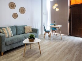 Una sala de estar moderna con un sofá verde, una mesa redonda de centro y una mesa de comedor con sillas. Se observan decoraciones en la pared y una puerta de madera al fondo.