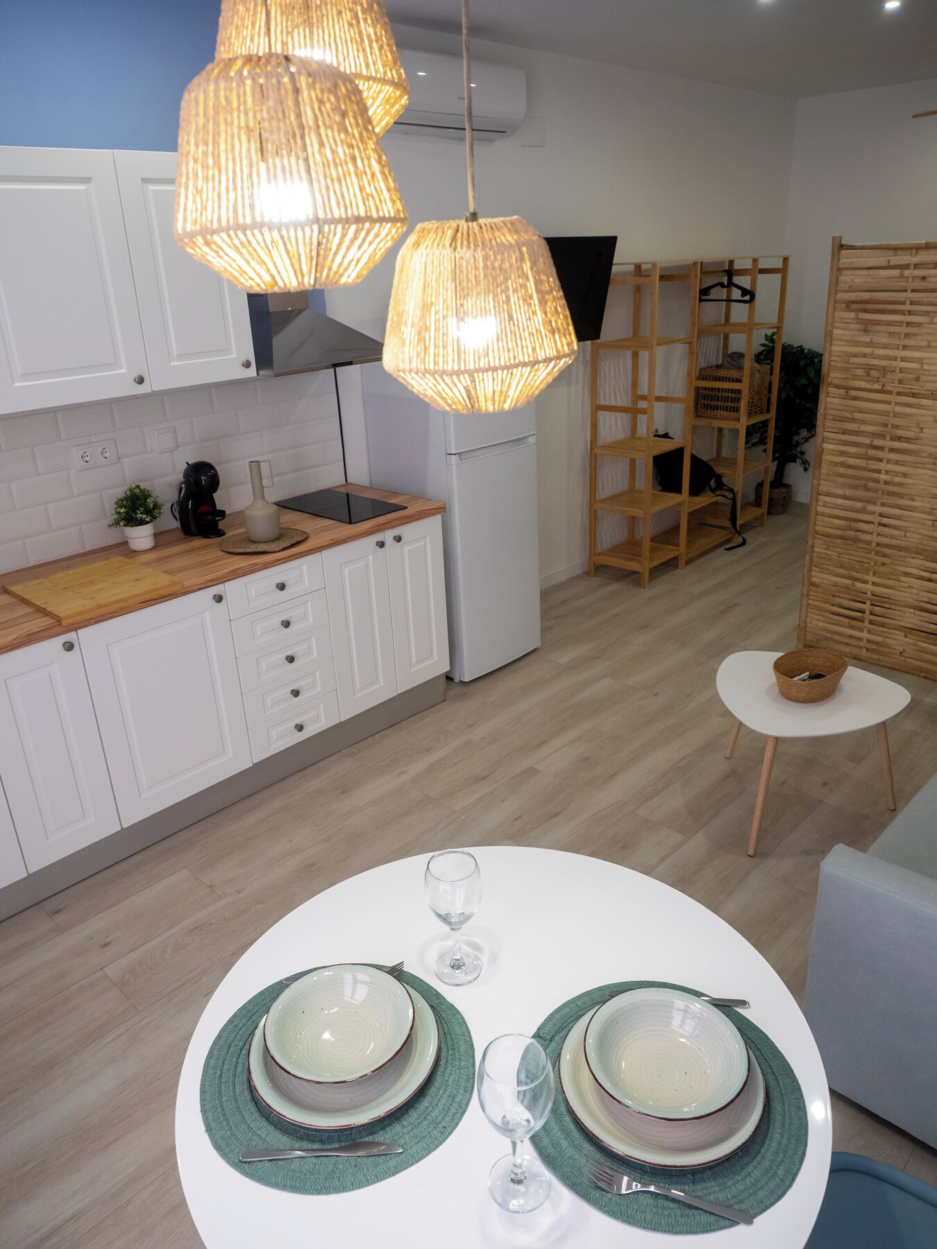 A modern kitchen with white cabinets, a white refrigerator, and a wooden countertop. There are two hanging wicker lamps, a small round table with a white tablecloth, and a dining area set with green placemats and white plates.
