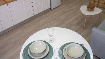 A modern kitchen with white cabinets, a white refrigerator, and a wooden countertop. There are two hanging wicker lamps, a small round table with a white tablecloth, and a dining area set with green placemats and white plates.
