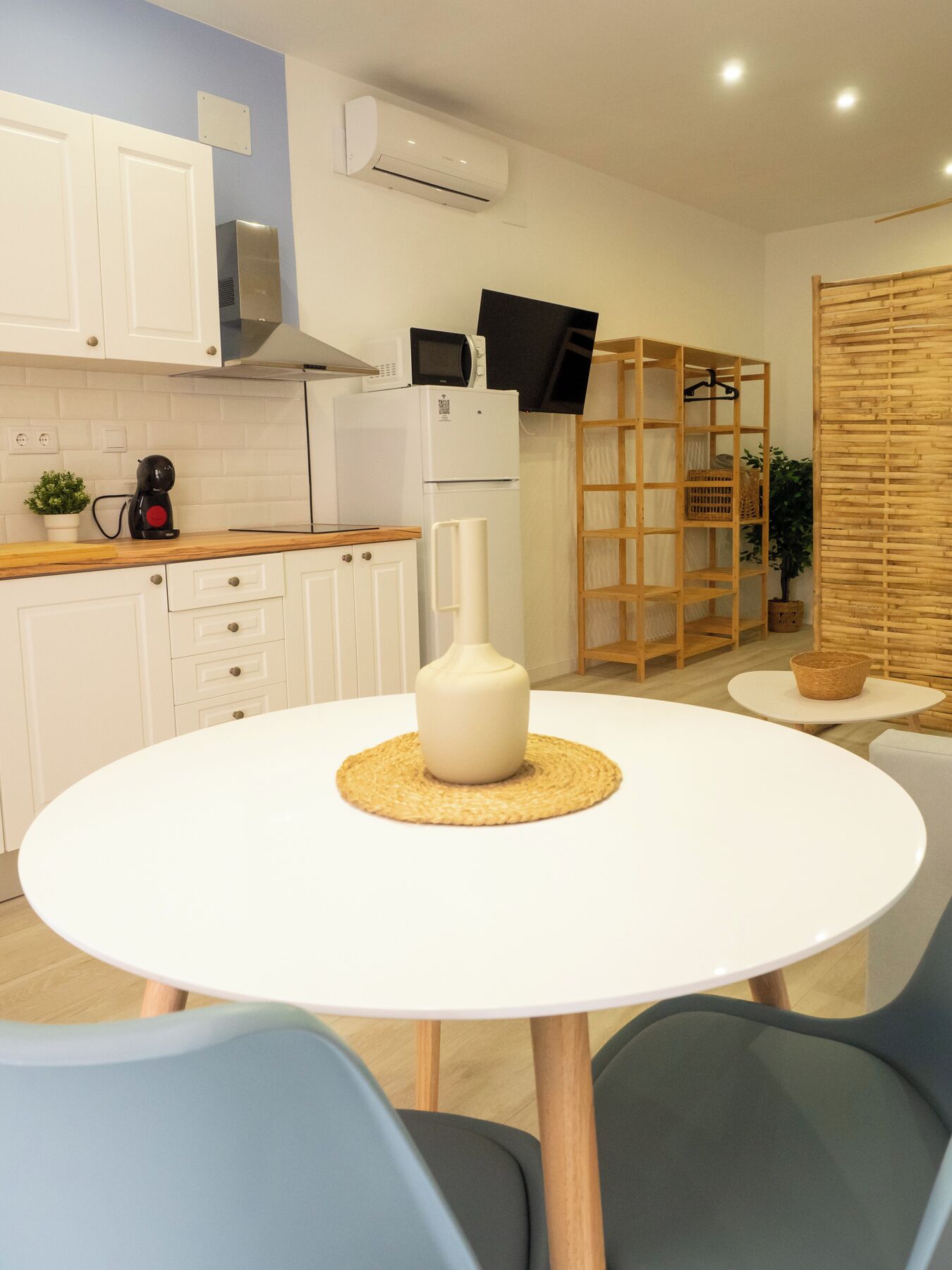 Una cocina moderna con armarios blancos, un microondas y una cafetera sobre la encimera. Hay una mesa redonda blanca con un mantel tejido y un florero.
