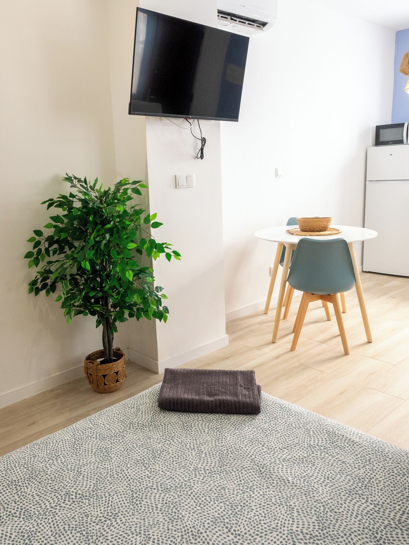 Ein modernes Wohnzimmer mit einem an der Wand befestigten Flachbildfernseher, einem Blumentopf, einem kleinen runden Tisch mit einem gewebten Korb und einem grauen Teppich.