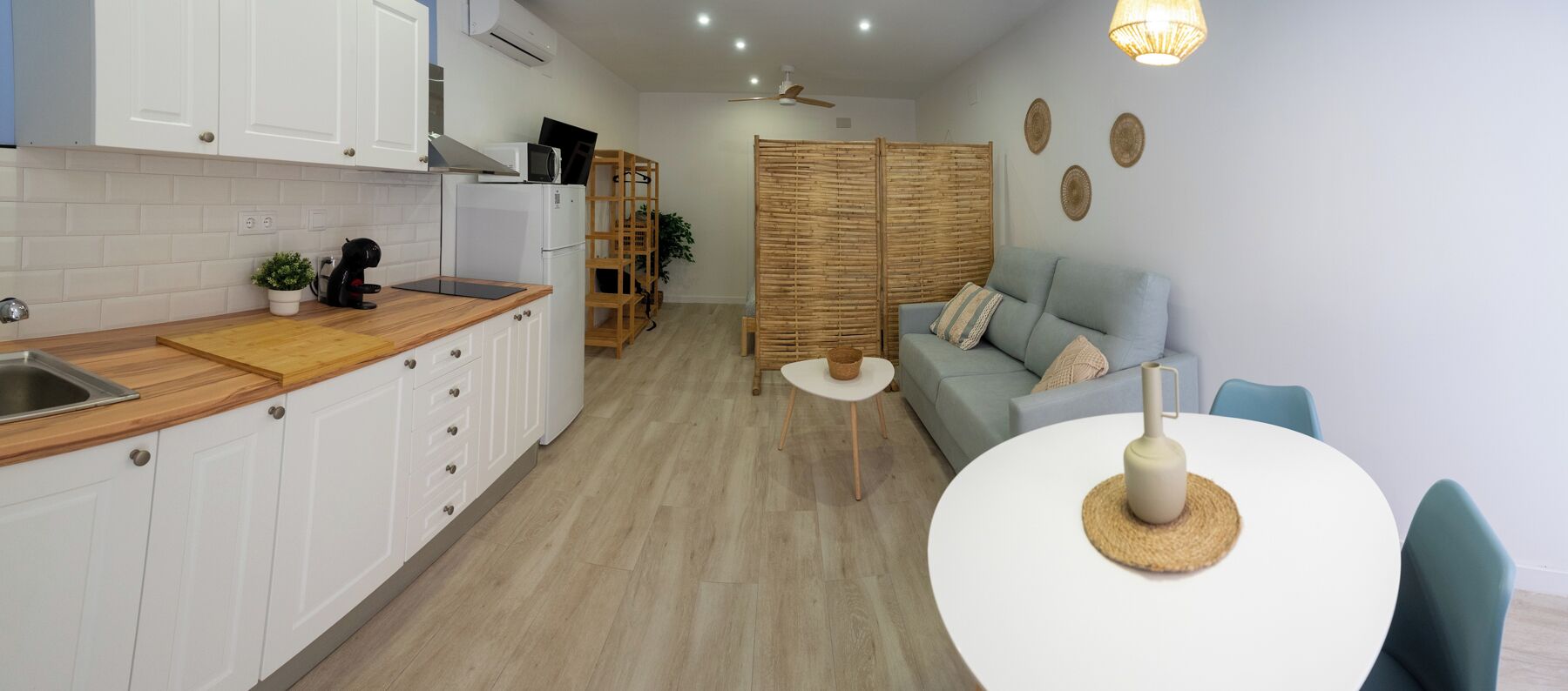 A compact kitchen with white cabinets, a wooden countertop, and a white refrigerator. There is a round table with a woven placemat and a vase. A light blue sofa is against the wall, and a bamboo screen divides the kitchen from a living area.