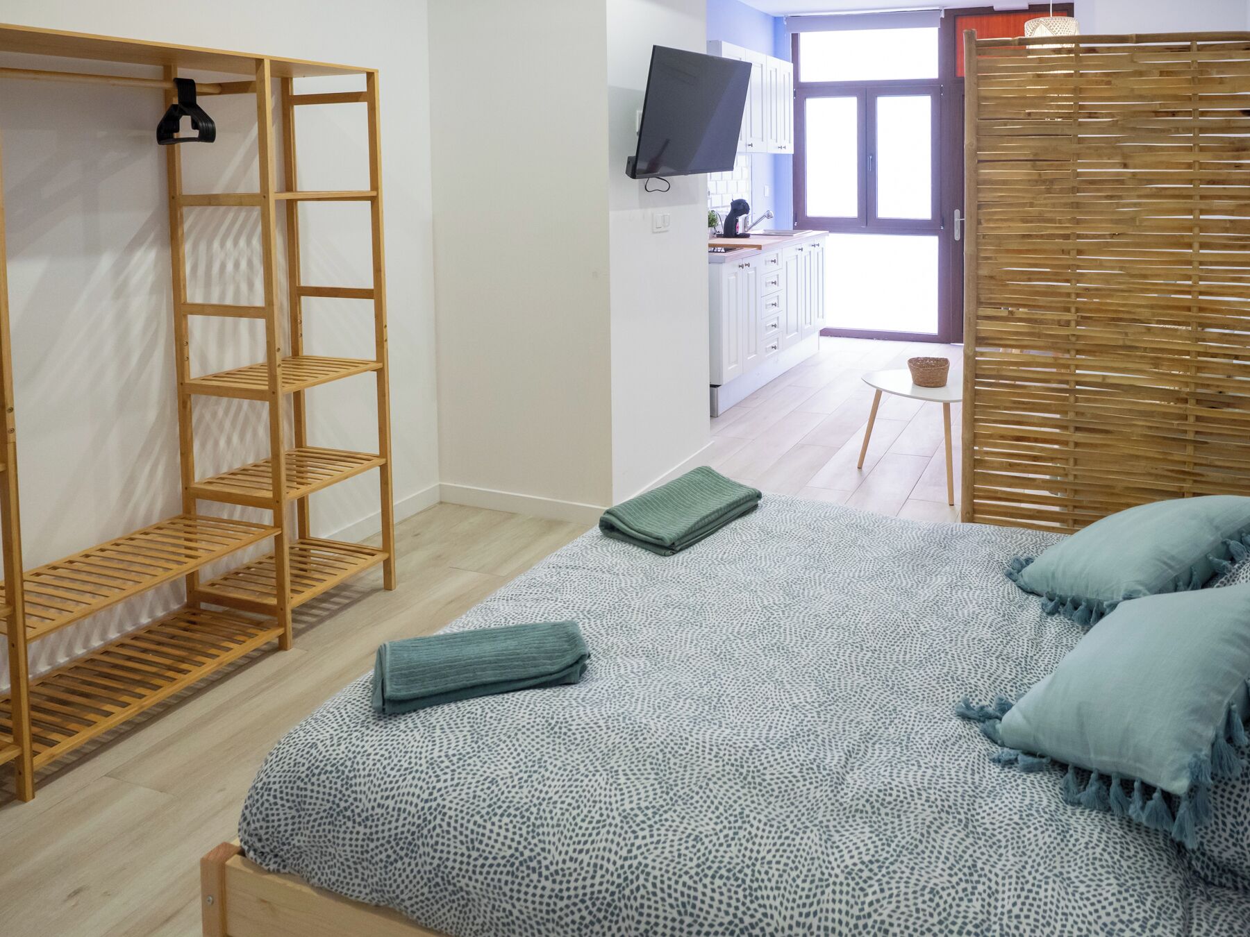 A bedroom with a bed, a wooden wardrobe, a television, and a kitchen area in the background.