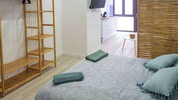 A bedroom with a bed, a wooden wardrobe, a television, and a kitchen area in the background.
