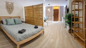 A bedroom with a wooden bed, a small stool, a macrame wall hanging, a bamboo room divider, and a wooden shelf unit. - Boho Beach Flats (València)